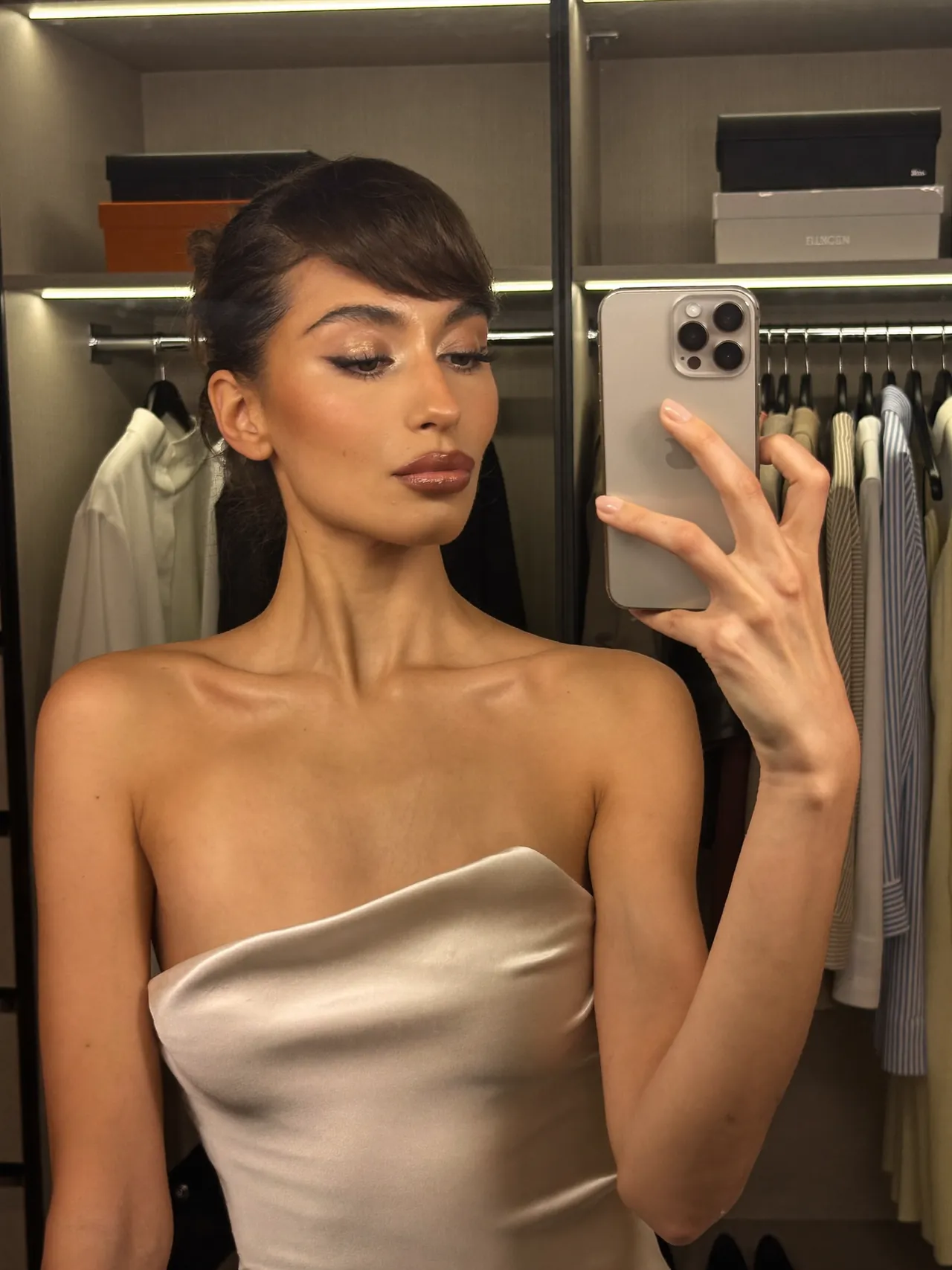 Une jeune femme se filme dans le miroir de sa chambre avec son téléphone, la lumière du jour derrière elle.
