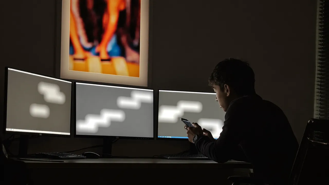 Un homme travaille de nuit sur un ordinateur portable, le visage éclairé par l'écran, un téléphone posé à côté.